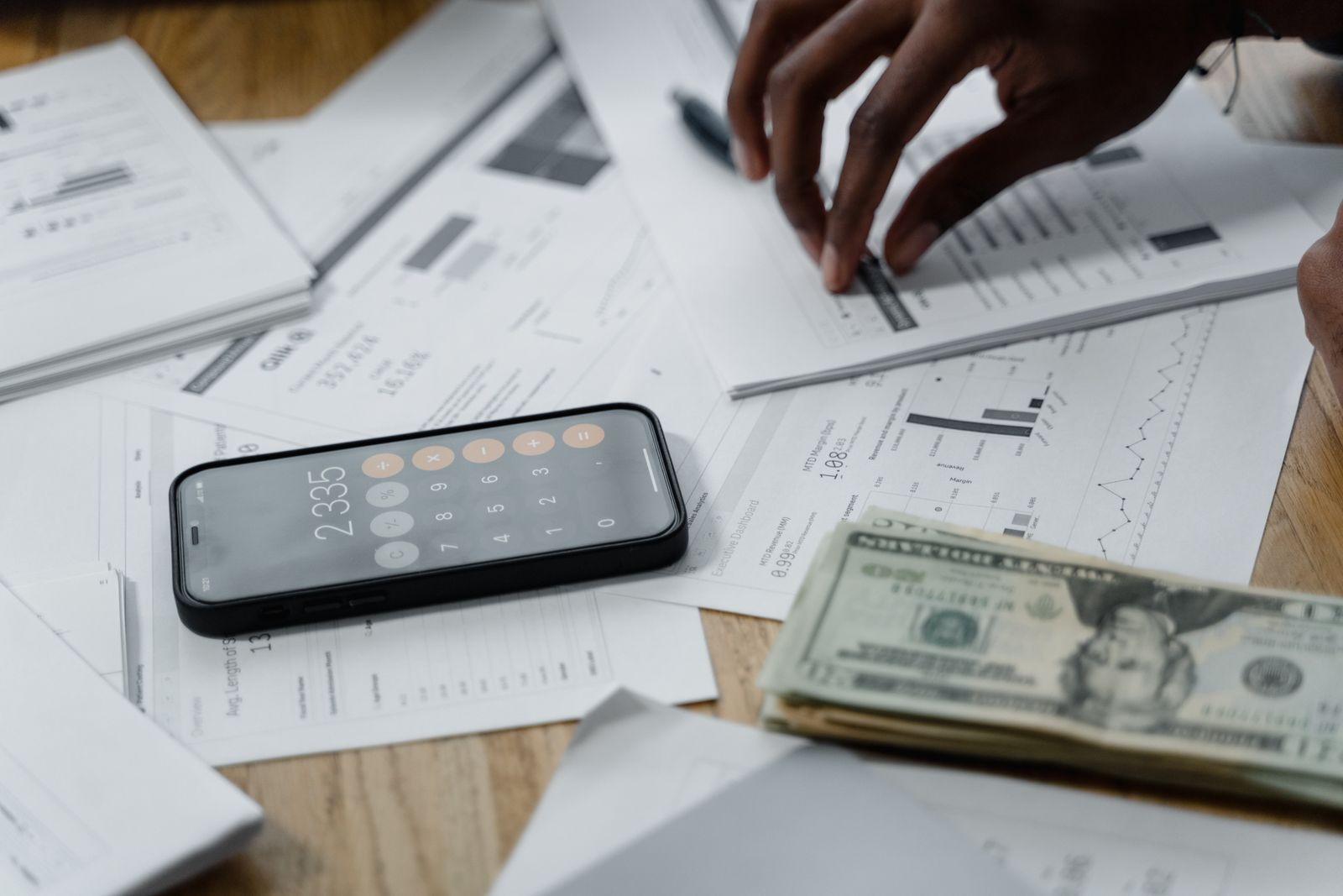 A phone calculator, cash, and financial reports spread across a desk with hands reaching for papers