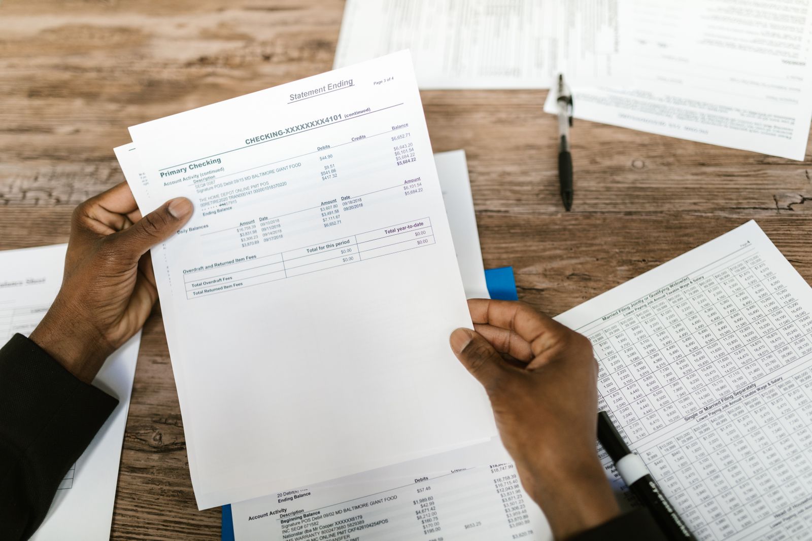 Hands holding a primary checking account statement over a desk covered in financial papers