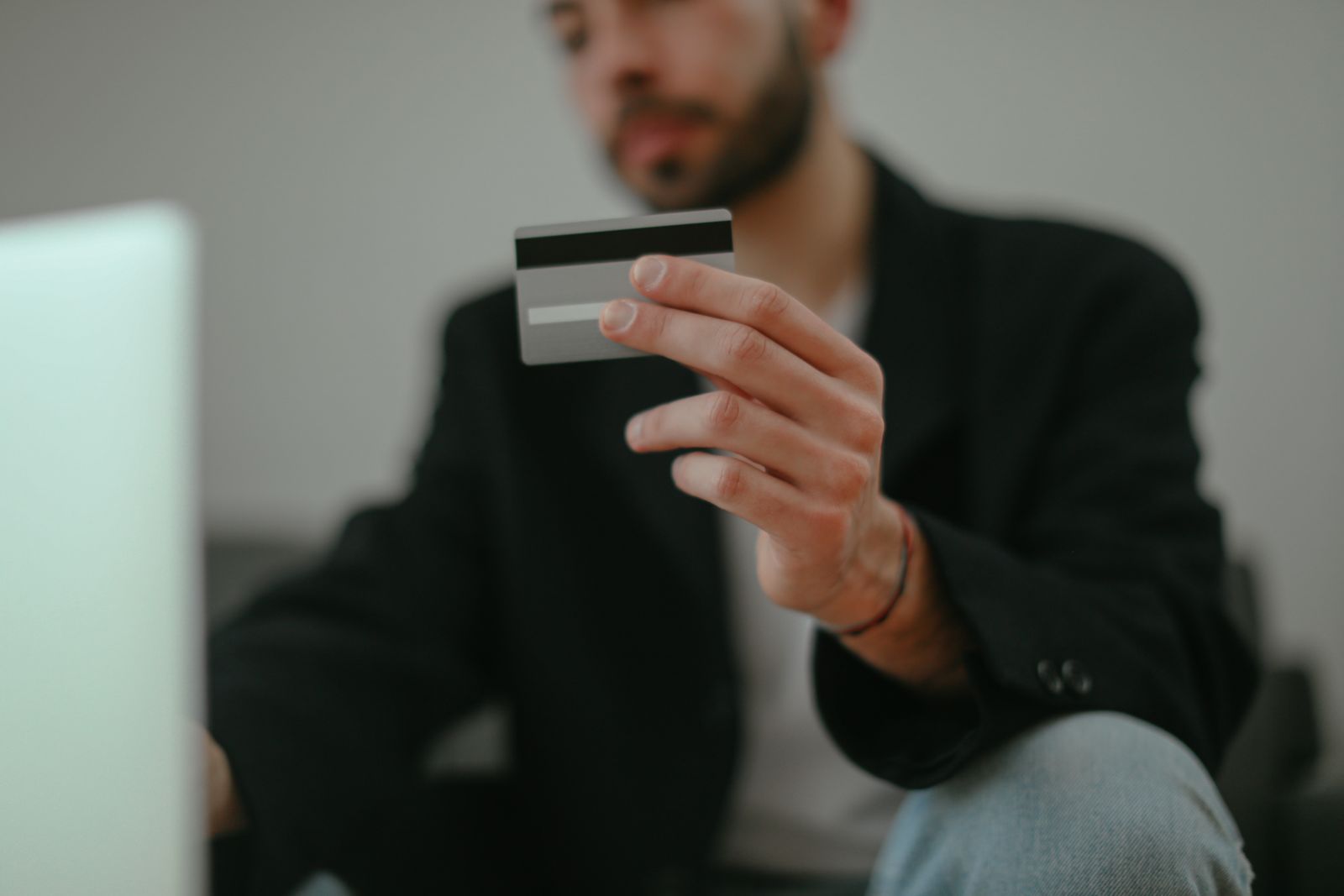 A young man with a blurred face holding a blank credit card in front of a laptop