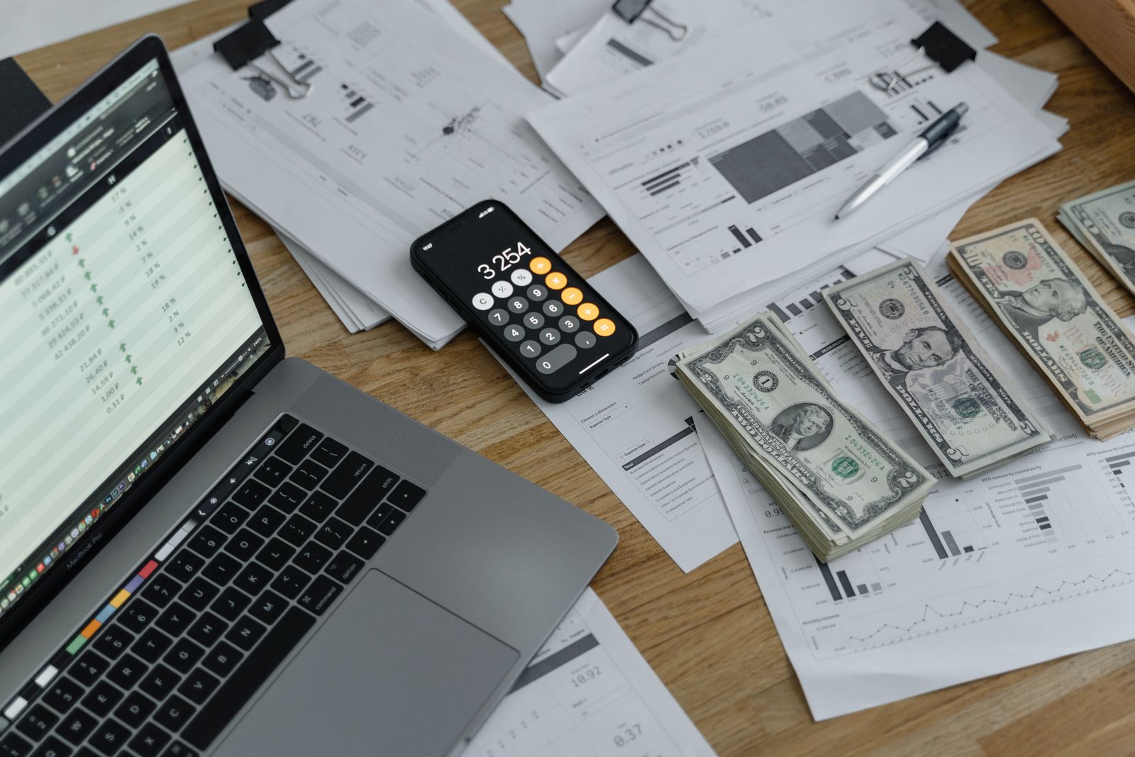 A laptop, phone calculator, cash, and paperwork spread across a wooden desk