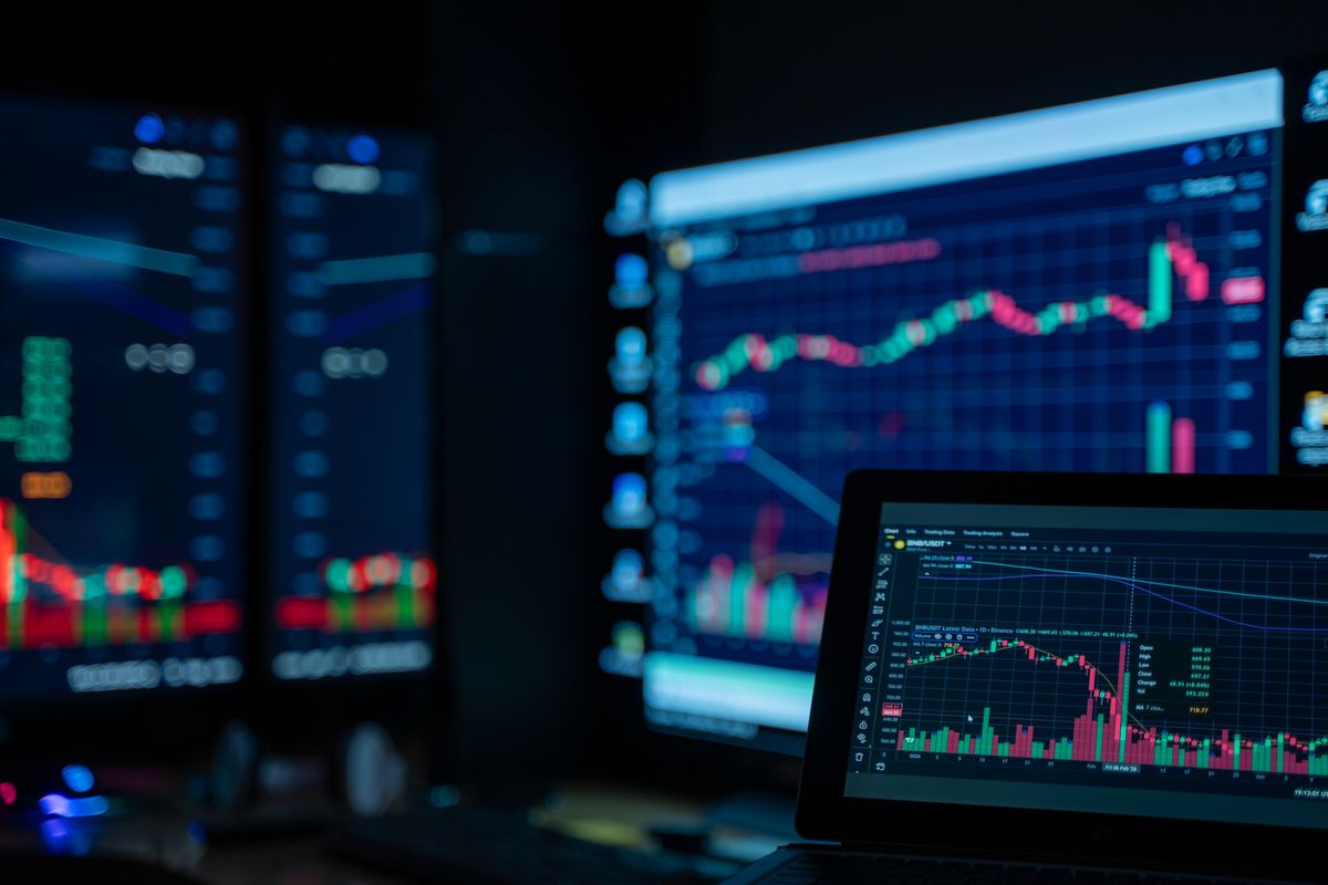 Several monitors in a dark room showing candlestick price charts