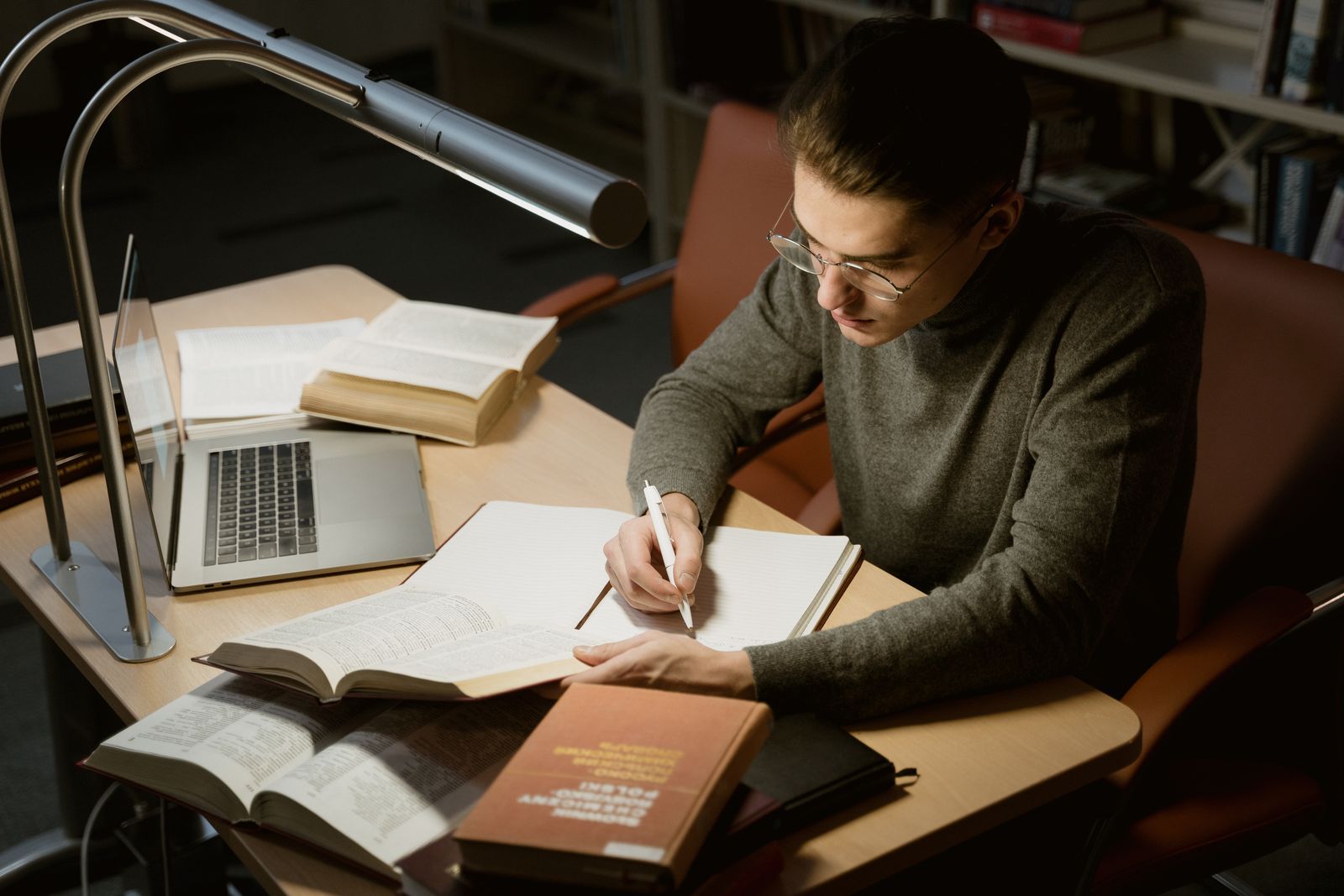 A young man in glasses writing in a notebook surrounded by open books at a library desk