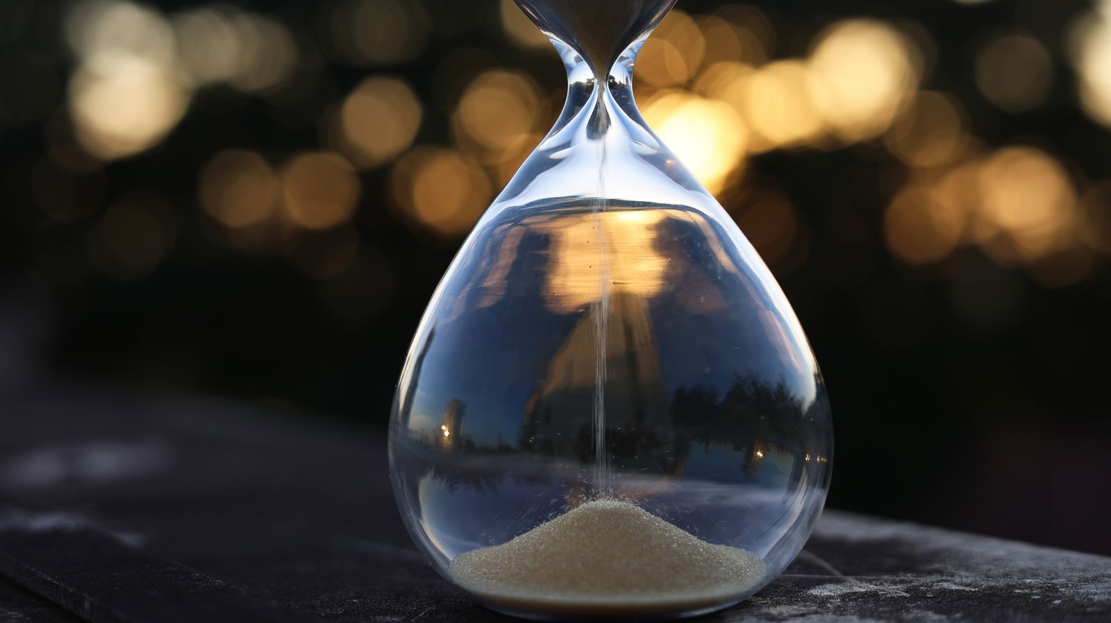 An hourglass with sand flowing through it silhouetted against warm bokeh lights at dusk