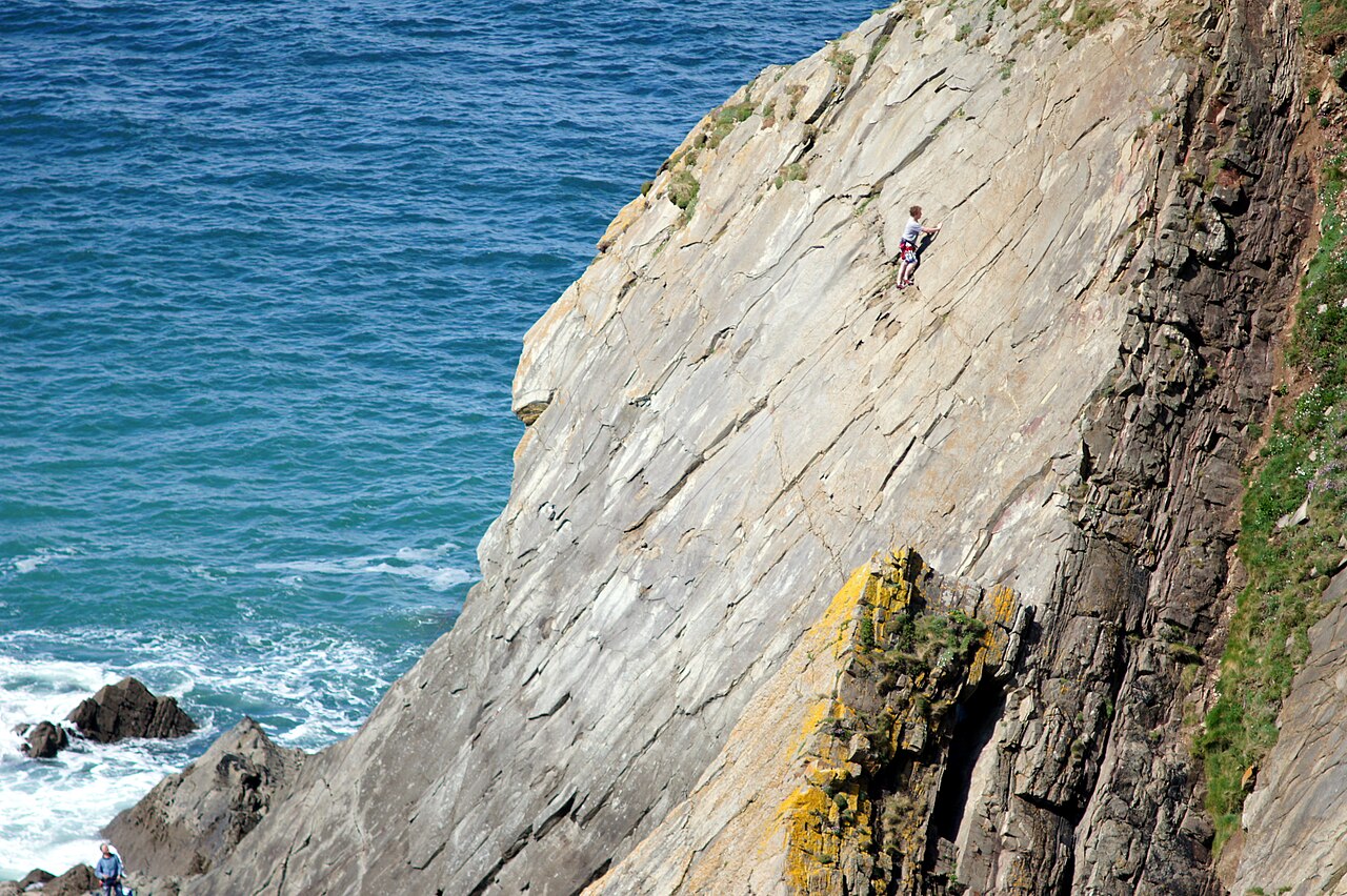 A lone climber partway up a steep sea cliff above open water