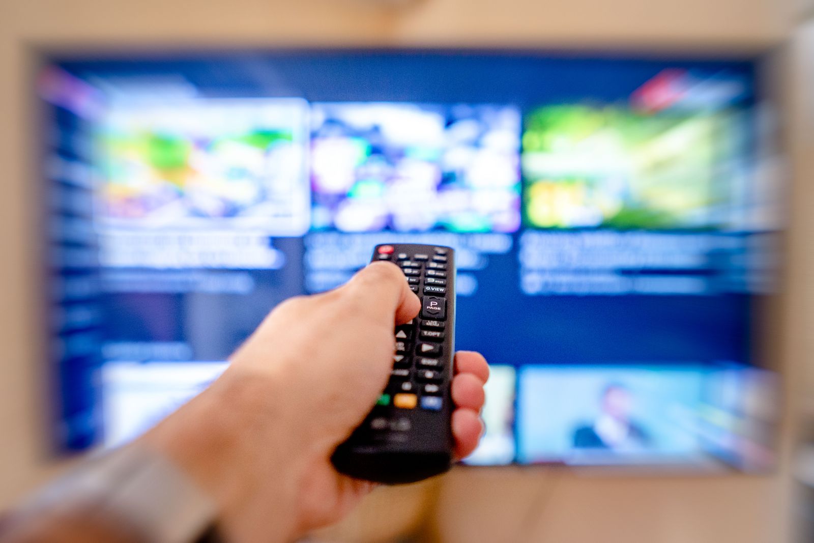 A hand holding a remote control pointed at a blurred television screen